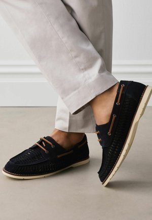 Black woven leather loafers with brown laces worn with light beige pants against a plain floor and wall background.