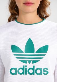 Sweat-shirt blanc avec un motif texturé, un logo Adidas vert et des accents verts autour du col.