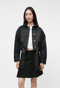 Chaqueta de mezclilla oscura, cropped, con grandes bolsillos, cierres de botón y un lazo en la cintura; llevada sobre una blusa blanca y una falda negra.