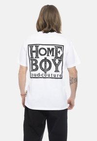 White short-sleeve T-shirt with black graphic text “HOME BOY loud-couture” on the back, featuring bold letters and a rectangular outline.