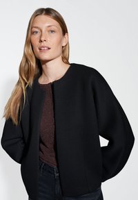 Giacca nera strutturata con maniche larghe, realizzata in un tessuto liscio. Indossata sopra un top bordeaux testurizzato con bottoni.