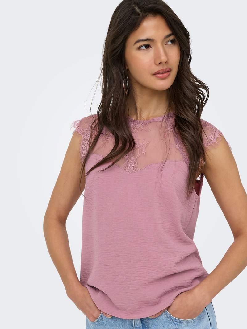 Jeune femme aux longs cheveux foncés portant un haut sans manches rose poudré avec des détails en dentelle, mains dans les poches, regard légèrement vers la droite.