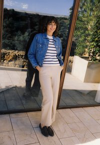 Frau, die draußen lächelt, trägt eine blaue Jeansjacke, einen gestreiften Pullover, beige Hose und dunkle Loafers, lehnt sich mit den Händen in den Taschen an eine Glastür.