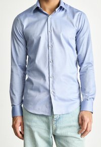 Homme portant une chemise bleu clair à manches longues boutonnée avec un col pointu et un jean bleu clair, debout devant un fond uni.