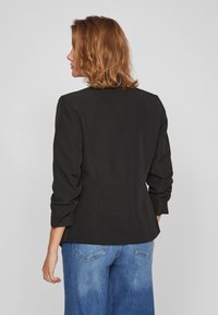 Blazer preto ajustado com um design estruturado, apresentando mangas franzidas e uma textura suave, combinado com jeans de perna larga em azul desbotado.