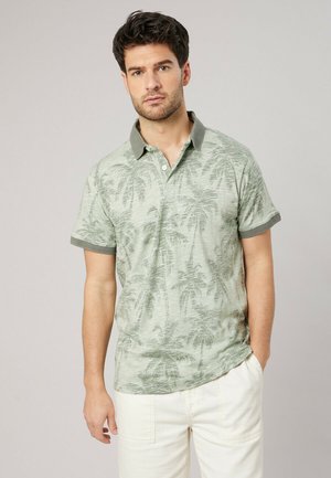 Homme portant un polo à manches courtes vert clair avec imprimé de palmiers et un pantalon blanc, debout devant un fond clair uni.