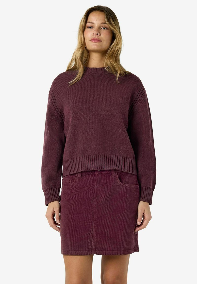 Femme portant un pull en laine violet foncé assorti et une jupe en velours côtelé, debout avec les mains relâchées le long du corps sur un fond blanc.