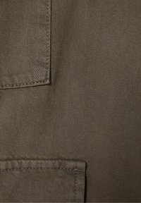 La tela de algodón en verde oscuro presenta una superficie texturizada con un detalle de bolsillo y bordes cosidos, lo que proporciona una apariencia estructurada.