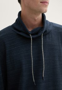 Dunkelblauer Hoodie mit hohem Kragen, ausgestattet mit einem strukturierten Muster und Kordeln mit mehrfarbigen Akzenten. Weicher Stoff und lockere Passform.