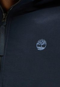 Mörkblå zip-jacka med en framträdande broderad logo av ett träd i ljusblått. Tillverkad av ett smidigt och lätt material.