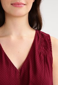 Burgundy top with a V-neckline, textured fabric, and floral embroidery on the left shoulder. Delicate bead detailing along the neckline.