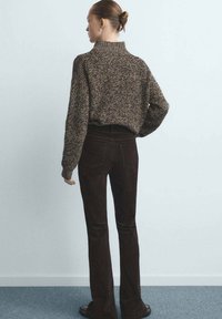 Stevige bruine gebreide trui met hoge hals, gecombineerd met donkerbruine wijde ribfluwelen broek. Model staat met de rug naar de camera voor een effen muur.