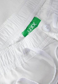 Balti šortai su elastinga liemens juosta, raišteliu ir sagos uždarymu. Žalia etikete su "JJXX" ženklu matoma šalia siūlės. Lygus paviršius.