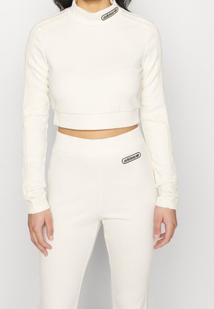 Nainen pukeutuneena valkoiseen Adidasin ribbineulottuun, lyhennettyyn pitkähihnaiseen topiin ja yhteen sopiviin korkeavyötäröisiin leggingseihin, joissa on mustat Adidas-logot kaulassa ja vyötäröllä.
