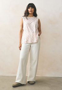 Világos rózsaszín mintás ujjatlan felső laza szabással, krémszínű bő szárú nadrággal és szürke klumpákkal párosítva, lágy, semleges háttér előtt.