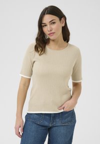 Top en maille côtelée beige avec des manches courtes, un col rond et des accents blancs aux poignets et à l'ourlet, associé à un jean en denim bleu.
