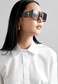 Sunglasses with oversized black frames, gradient lenses, and gold accent hardware. Worn with a crisp white button-up shirt and gold earrings.