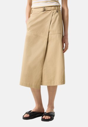 Beige Wickel-Midirock mit Gürtel, kombiniert mit schwarzen Slip-On-Sandalen und einem gestreiften Oberteil, getragen von einer stehenden Person.