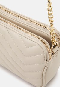 Beige kviltet skinn crossover veske med gullhardware, som har to glidelåslommer og en kjede-rem. Glatt tekstur og rene linjer.
