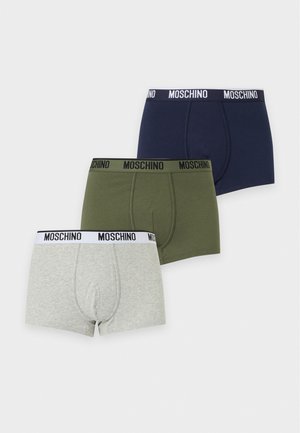 Un mannequin masculin se tient devant un fond blanc uni, vêtu d’un boxer Moschino bleu marine.