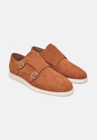 Scarpe in suede marrone con una superficie liscia, dotate di due cinturini regolabili e suola in gomma chiara. Design elegante per un facile indossamento.
