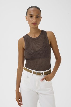 Femme porte un haut marron sans manches en maille côtelée, rentré dans un pantalon blanc taille haute avec une ceinture marron, debout devant un fond clair uni.