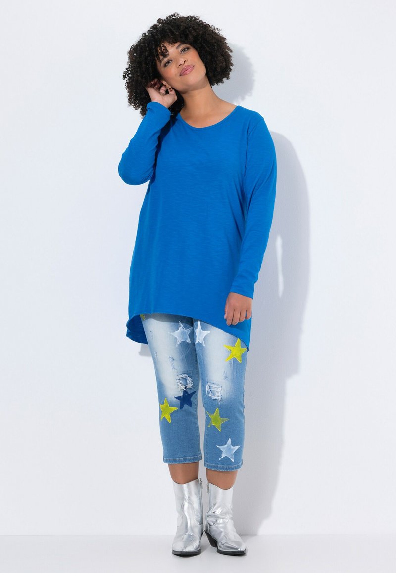 Blaues langärmeliges Tunika-Shirt kombiniert mit ausgewaschenen hellblauen Jeans mit Sternmustern in Gelb und Weiß, ergänzt durch silberne Ankle Boots.