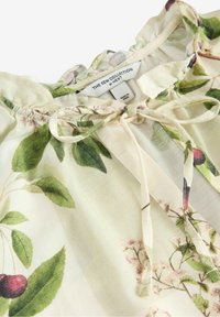 Květinová blůza z lehkého materiálu se světlým slonovinovým pozadím. Obsahuje vzory zelených listů a třešní, nastavitelné zavazování u krku a volánkový límec.