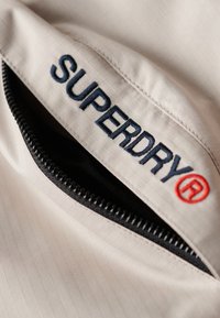 Beige jakke stof med gitterstruktur, med en sort lynlås. "SUPERDRY" er broderet i mørkeblå med en rød cirkulær logoaccent.