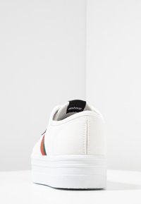 Baskets en toile blanches avec une finition texturée, rayures contrastées vertes et rouges, et une semelle en caoutchouc blanc solide. Étiquette de logo noire à l'arrière.