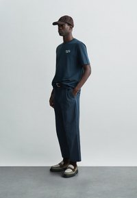 Muži vo námorníckej modrej tričko, zodpovedajúcich nohaviciach, hnedou čiapkou, tmavými ponožkami a dvojfarebnými mokasínami stoja na pozadí s jednoduchou šedou farbou.