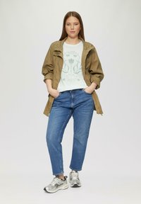 Olijfgroene lichtgewicht jas, witte grafische t-shirt, blauwe spijkerbroek en zilveren sneakers. Model poseert met handen in de zakken.