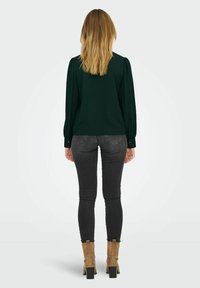Dunkelgrüne Bluse mit langen Ärmeln und gepufften Schultern, kombiniert mit enganliegenden grauen Jeans und beigen Ankle Boots mit Blockabsatz.