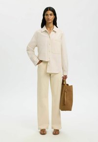 Femme debout portant une chemise ample couleur crème et un pantalon large assorti, tenant un sac fourre-tout en daim marron dans sa main droite.
