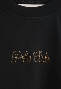 Camiseta negra de cuello redondo con "Polo Club" en letras doradas con tachuelas. La tela es lisa y elástica con un color sólido.