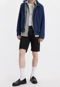Chaqueta de bombardero azul marino sobre una camiseta gris, pantalones cortos de mezclilla negros, calcetines blancos y mocasines negros. Patrón de cuadros notable en el interior de la chaqueta.