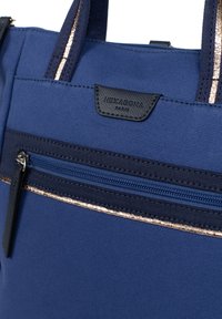 Borsa tote in tela blu con tasca frontale con zip, patch in pelle nera con logo e sottili accenti metallici lungo la parte superiore.