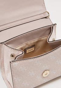 Beżowa torebka Guess z białym wytłaczanym wzorem, otwartą klapą, beżowym materiałem wykończeniowym w środku oraz widocznym metalowym zapięciem na zatrzask.