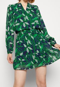 Kukkapainettu mekko, jossa on tummansininen tausta, vihreitä lehtiä ja vaaleita yksityiskohtia, pitkät hihat, joustava vyötärö ja kerroksellinen hame.