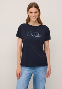 Leende kvinna klädd i en marinblå t-shirt med texten "Vandra och förbli nyfiken" över ljusblå jeans, mot en neutral bakgrund.