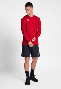 Rød langærmet atletisk skjorte med rund hals og logo, parret med sorte shorts. Modellen har sorte sko og sokker på og har armene korslagte.