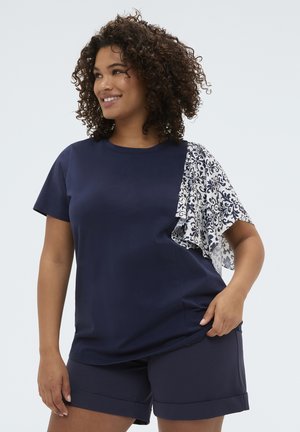 Uśmiechnięta kobieta o kręconych włosach nosi granatowy top z falbanianym rękawem w czarno-biały wzór oraz pasujące granatowe szorty.