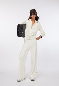 Donna con cardigan bianco con cerniera e pantaloni a gamba larga che tiene una grande borsa imbottita nera, indossa occhiali da sole neri e sneakers bianche.