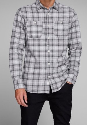 Mann trägt ein graues und schwarzes kariertes Hemd mit langen Ärmeln und zwei Brusttaschen vorne, kombiniert mit einer schwarzen Hose.