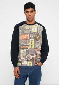 Maglione lavorato a maglia con un design grafico caratterizzato da toppe multicolori a tema videoludico. Maniche di un blu scuro e scollatura a girocollo a costine.