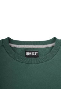 Grön sweatshirt med en texturerad ribbad halsringning, med en grå inredning. Svart etikett som säger "ÄRLIGHET ÄR REGELN" vid kragen.