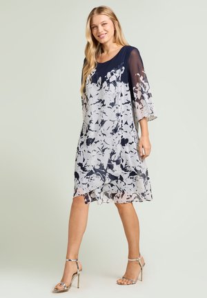 Femme blonde portant une robe fleurie bleu marine et blanche longueur genou avec manches transparentes et sandales argentées à talons hauts sur un fond uni.