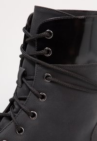 Svart läderstövel med glansiga detaljer, med snören genom silverfärgade öljetter och en texturerad yta. Hög design med förstärkta sömmar.