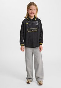 Svart långärmad skjorta med gula accenter och logotyp, i kombination med gråa sweatpants. Skjortan har en kragliknande hals och elastiska ärmslut.