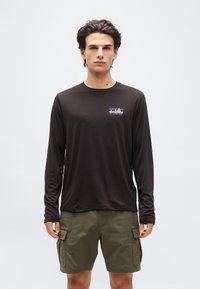 Νεαρός άνδρας που κοιτάζει μπροστά, φοράει μαύρο μακρυμάνικο πουκάμισο Patagonia και χακί cargo σορτς, μπροστά σε ένα απλό λευκό φόντο.